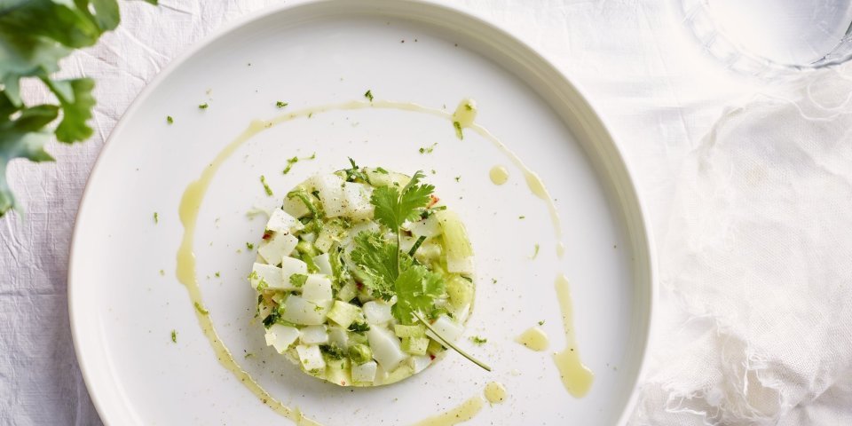 Tartaar van zeekat met komkommer, lente-ui, koriander en chilipeper Op tafel ligt een mooi wit linnen tafelkleed, waarop het witte bord staat. Hierop ligt de tartaar van zeekat met komkommerstukjes, lente-ui en koriander. We voegden ook wat chili toe voor extra pit en als afwerking nog wat olijfolie en een korianderblaadje.