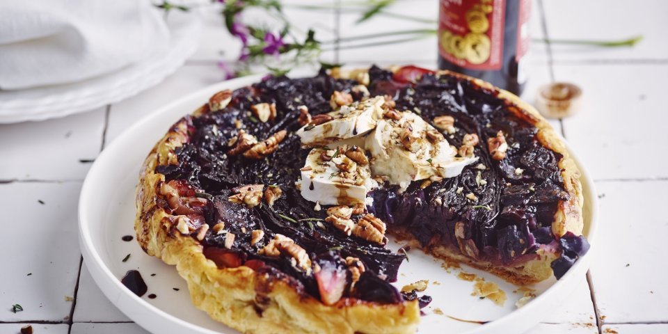 Een tarte tatin van rode kool met geitenkaas ligt op een groot wit bord. Er is al een stukje aangesneden en ze werd afgewerkt met enkele druppels balsamico en pecannoten. Achteraan zie je de fles balsamico nog op het aanrecht staan alsook een paars bloemetje. 