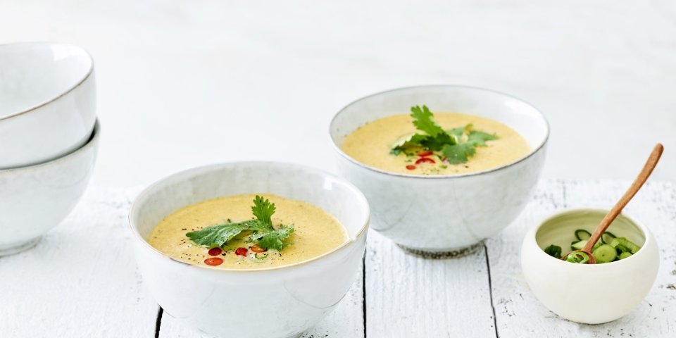 Thaise tomatensoep in twee kommetjes uitgeschonken. Topping van chilipeper en koriander ligt mooi in het midden van de soep. Alles staat op een witte houte tafel.