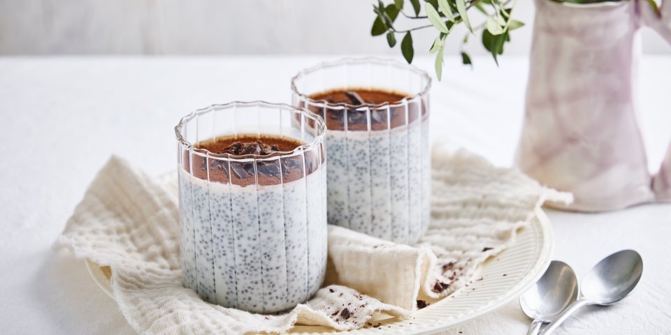 Tiramisu chiapudding met chocolade Op een bord staan twee grote glazen gevuld met de chiapudding, hij is afgewerkt bovenaan met cacaopoeder en chocoladeschilfers. Er liggen twee zilvere lepeltjes langs.