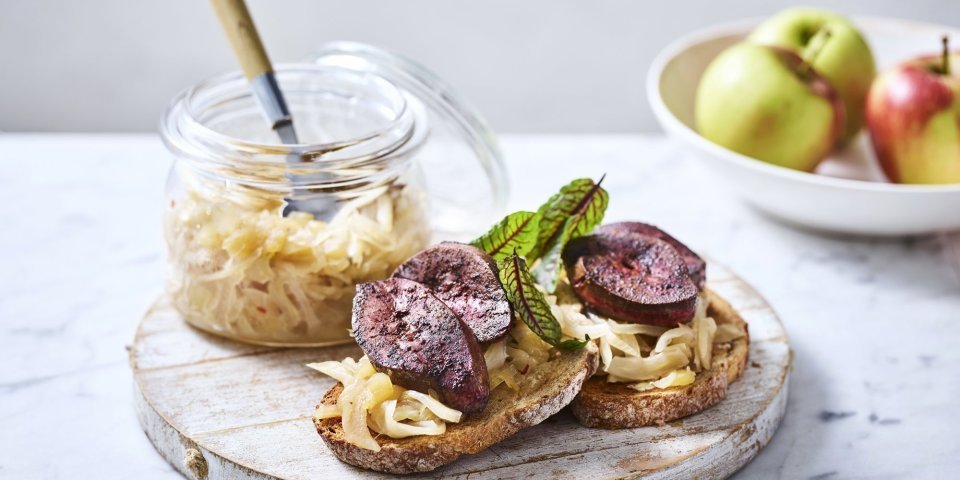 Twee toasten liggen op een houten serveerplank. Ze zijn belegt met chutney en gebakken niertjes. De overige chutney staat er langs in een glazen bokaal. 