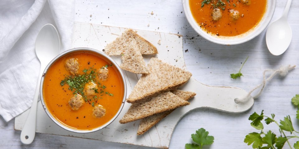 Heerlijk gemaakte tomatensoep staat uitgeschenkt op tafel in twee soepborden. De balletjes drijven in de soep en is afgewerkt met peterselie. Brood en lepels liggen langs het bord, klaar om te slurpen.