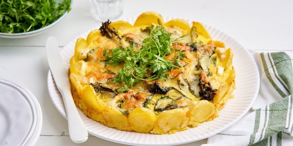 De tortilla ligt op een groot wit bord, er zijn aardappelschijfjes gebruikt als bodem maar ook als rand. Dit geeft een mooi beeld om naar te kijken. De taart is gevuld met prei, broccolini en spinazie, boordevol groenten. We serveren hem nog met wat kervel om het af te werken. 