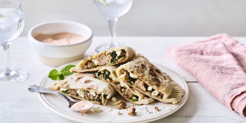 Turkse pannenkoek met gehakt, spinazie en feta De turkse pannenkoek gevuld met gehakt, spinazie en feta ligt op een wit bord. Er ligt een roze servette langs en het chili-dipsausje staat bovenaan.