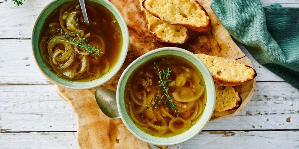 Twee kommetjes uiensoep staan uitgeschonken. Er liggen uiringen en verse tijm in de soep, kaasbroodjes liggen langs de soep. Klaar om te eten en warm te krijgen.