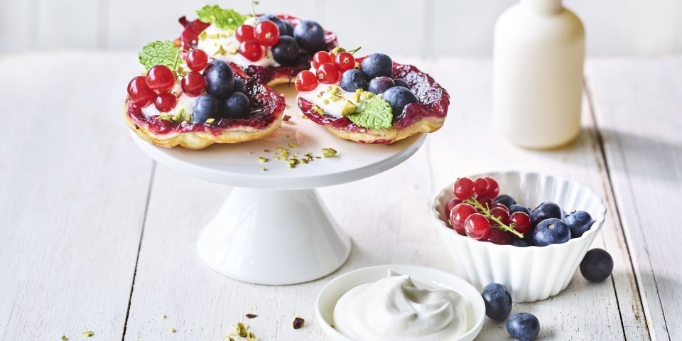 Upside down taartje met aalbessen en blauwe bessen Op een witte taartschaal liggen 3 kleine taartjes met zomerfruit. Hiernaast ligt nog wat opgeklopte room, nog een schaaltje met extra rood fruit en een vaasje met roze bloemetjes.