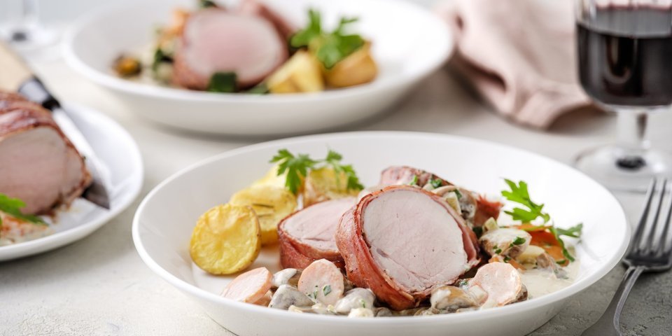 Varkenshaasje met champignonsaus en wortelen Op tafel staan twee porties uitgeschept. Het heerlijk bereide varkenshaasje ligt op de romige champignonsaus met plakjes wortel. Er liggen ook ovengeroosterde aardappeltjes bij. Langs het bord ligt een servette, glaasje rode wijn en bestek.