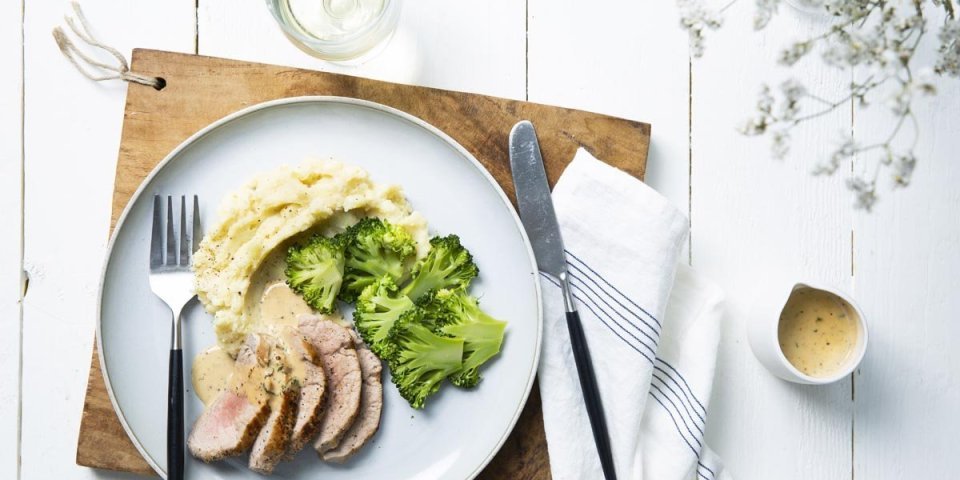 Varkenshaasje met mosterdsaus en puree Op tafel ligt een houten snijplank met hierop een lichtblauw groot plat bord. Hierop is het hoofdgerecht geserveerd, stukjes varkenshaasje in sneetjes, plakjes broccoli en een een schep aardappelpuree. Het sausje zit in een sauskannetje langsheen het bord.
