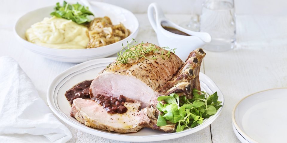 Een varkenskroontje ligt op een groot wit bord, aangesneden en opgediend met de rode wijn saus. In de achtergrond staat de aardappelpuree en de stukjes witloof. Het varkensvlees is mooi roos gebakken. 
