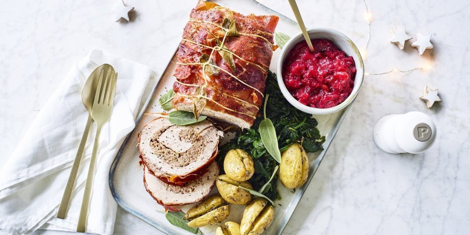 Varkensrollade met gehakt, veenbessencompote en krieltjes uit de oven Een mooi aangesneden varkensrollade ligt op een rechthoekig dienblad. De aardappeltjes werden net niet doormidden gesneden alvorens ze de oven in gingen met een kruidentakje. De veenbessencompote staat langs de rollade die zorgt voor een intense kleur in deze feestmaaltijd. Goud bestek ligt langs het gerecht.