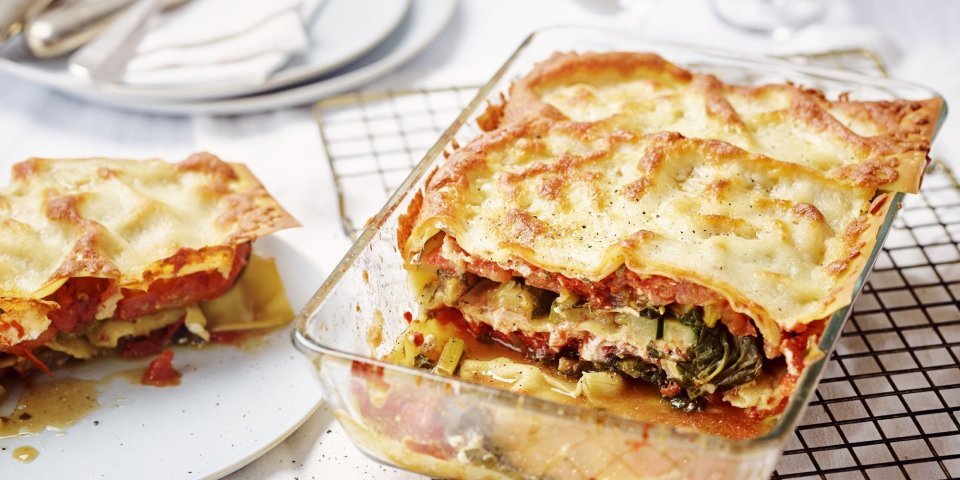 Vegetarische lasagne met snijbiet, courgette en ricotta De lasagne staat op tafel in een glazen doorzichtige ovenschotel. Hij is aangesneden zodat je mooi de laagjes ziet van pasta, groenten en saus. De portie die is uitgeschept ligt op een bordje er naast.