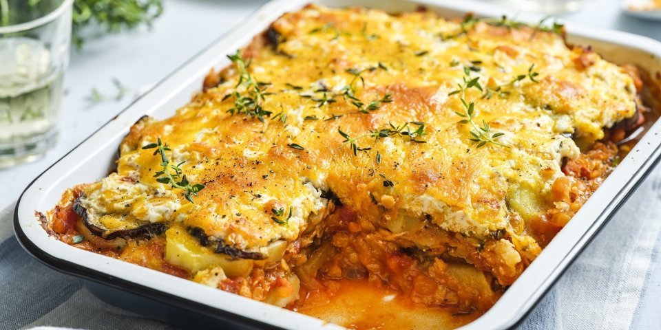 Vegetarische moussaka met linzen Er staat een grote schotel met vegetarische moussaka op tafel. Er is al een portie uitgeschept en op een bordje geserveerd. De moussaka is afgewerkt met verse tijmblaadjes. Op tafel staan ook nog 2 glazen en het plantje verse tijm.