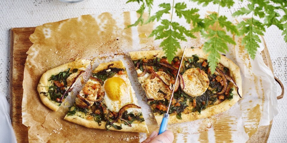 Vegetarische pide met spinazie en geitenkaas Een hartige Turkse groententaart met spinazie, geitenkaas en ei ligt op een houten snijdplank. Ze wordt aangesneden met een mes.