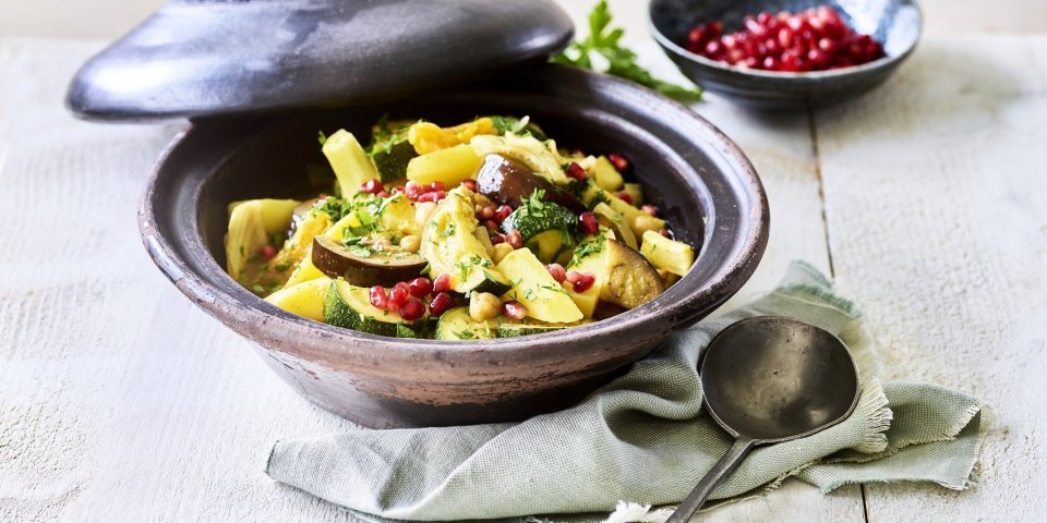 Vegetarische tajine Er staat een tajine op tafel, de tajine heeft een zwarte kleur en is gevuld met stukjes aardappel, courgette, venkel, abrikozen, kikkererwten en granaatappelpitten.