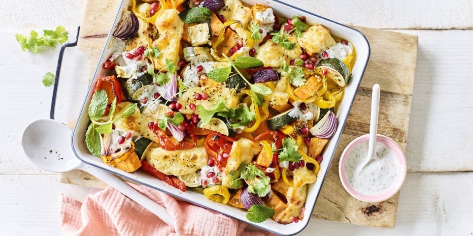 Vegetarische traybake met aardappel, rode ui, paprika en Berloumi Op een witte tafel staat een houten snijplank met hierop een vierkante ovenschotel. Hij is gevuld met zoete aardappel, courgette, paprika en rode ui. Er is ook Berloumi gebruikt in dit gerechtje.