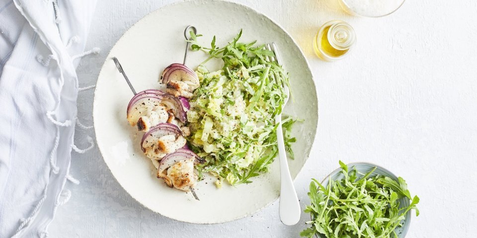 Visbrochette van hondshaai met groene puree Een brochette van vis met stukjes rode ui liggen op een wit rond bord, bovenop de groene puree.