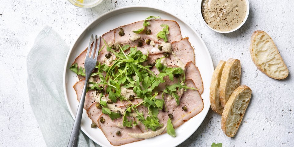 Vitello sgombro Deze vitello sgombro ligt op een wit bord, heerlijk afgewerkt met de koude saus, kappertjes en rucola. Er liggen ook enkele stukjes brood langs het bord.