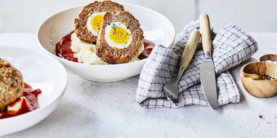 Vogelnestjes in tomatensaus met aardappelpuree In een diep wit bord ligt de aardappelpuree onderaan met verse rode tomatensaus en daar bovenop de doormidden gesneden vogelnestjes. Er ligt een handdoek langs met blauwe ruitjes, hierop het bestek met houten handvat.