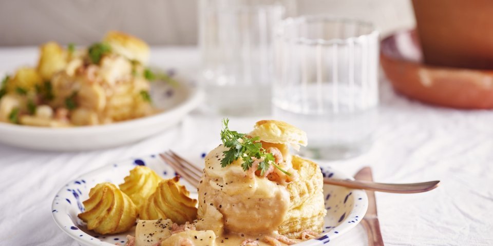 Vis vol-au-vent van schelvis en grijze garnalen Twee borden staan klaar om van te smullen. Vol-au-vent van vis in een bladerdeegpasteitje met pommes duchesse. Er staat een glaasje water naast het bord.