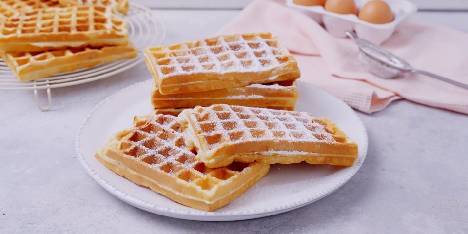 Wafels met Belgische peperkoek Een stappeltje klaargemaakte wafels ligt op een groot wit bord. Bloemsuiker is rijkelijk op de wafels gestrooid. In de achtergrond zie je nog enkele eitjes liggen en een roze linnen servette.