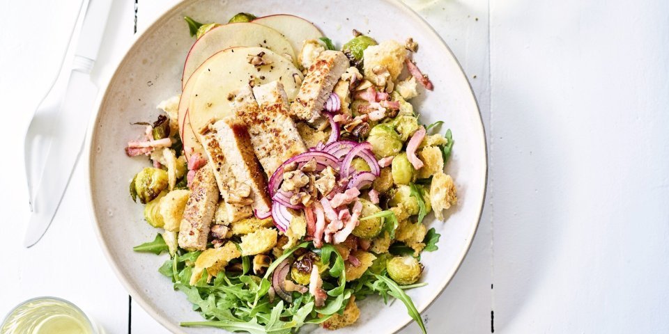 Winterse panzanella met ovengeroosterde spruitjes, versgesneden appel en gegrilde kalfsoester in een diep wit bord. Wit bestek en wijn staat langs het bord, klaar om aan te vallen.