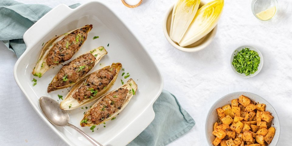 Witloof gevuld met gehakt en champignons met kruidige aardappelen Gevulde kroppen witloof met kruidig gehakt liggen in een ovenschotel. De schotel staat op een lichtblauwe doek met kruidige aardappelen in kommetje langs.