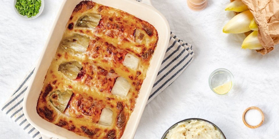 Gegratineerde witloofrolletjes opgerold in gekookte ham liggen mooi naast elkaar in een ovenschotel. De kaassaus is laat de rolletjes net bovenkomen. Er staat een pot heerlijke puree klaar om aan te vallen. 