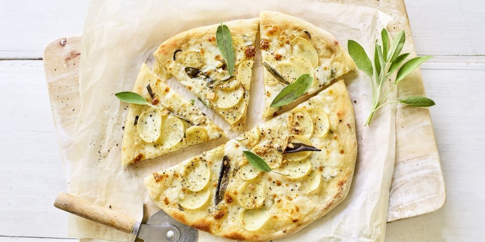 Witte pizza met aardappelplakjes, mozzarella en salie Een pizza belegt met mozzarella, plakjes aardappel en salie ligt op een wit boterpapiertje. Er ligt een pizzasnijder langs de pizza en nog enkele verse blaadjes salie.