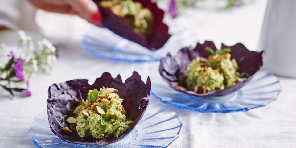 Wrap van rode kool met erwtenhummus en gerookte amandelen Er liggen 3 wraps van rode koolbladeren, elk op een blauw kristallen bordje. De wrap is gevuld met hummus van groene erten en geroosterde en gerookte amandelen. Een vrouwenhand neemt alvast een wrap tot haar hapje, geen tijd te verspillen dus.