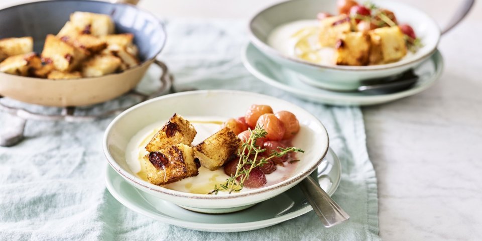 Een heerlijk én makkelijk ontbijtje staat klaar op tafel. De yoghurt is geserveerd in ontbijtkommetjes en afgewerkt met gegrilde druiven en krokante croutons van rozijnenbrood.