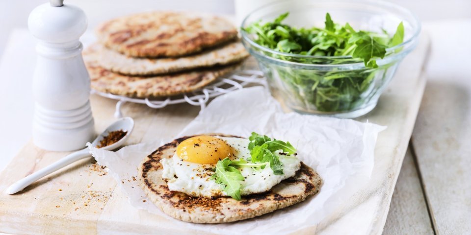 Yoghurtbrood van boekweitmeel met spiegeleitjes Het yoghurtbrood ligt op een houten snijplank. Hierop liggen meerdere ingrediënten om aan de slag mee te gaan, denk maar aan rucola, peper, extra yoghurt.