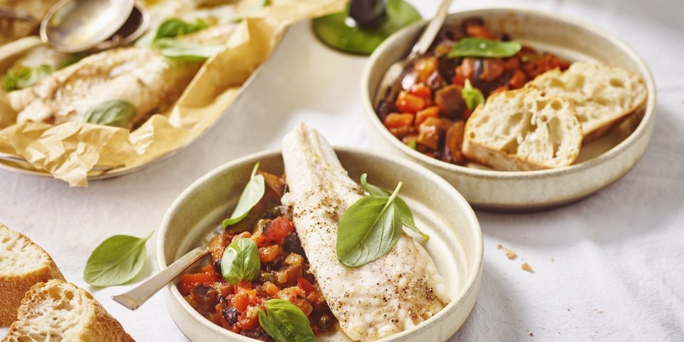 Zeeduivel uit de oven met caponata Twee bordjes gevuld met heerlijke homemade tomatensaus met olijfjes en kappertjes. Hier bovenop ligt het stukje vis en enkele blaadjes basilicum. Er staat ook een glaasje wijn op tafel met groene voet.