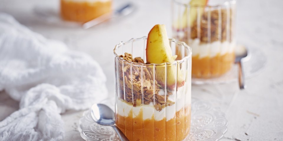 Zoete aardappel met kwark, granola en appel Op tafel staan 3 glaasjes gevuld met dit ontbijtje. Onderaan ligt een lepeltje zoete aardappel, hierbovenop de kwark, granola en een stukje gestoofd appel. De kleuren van dit ontbijtglaasje zijn gewoon om blij van te worden.