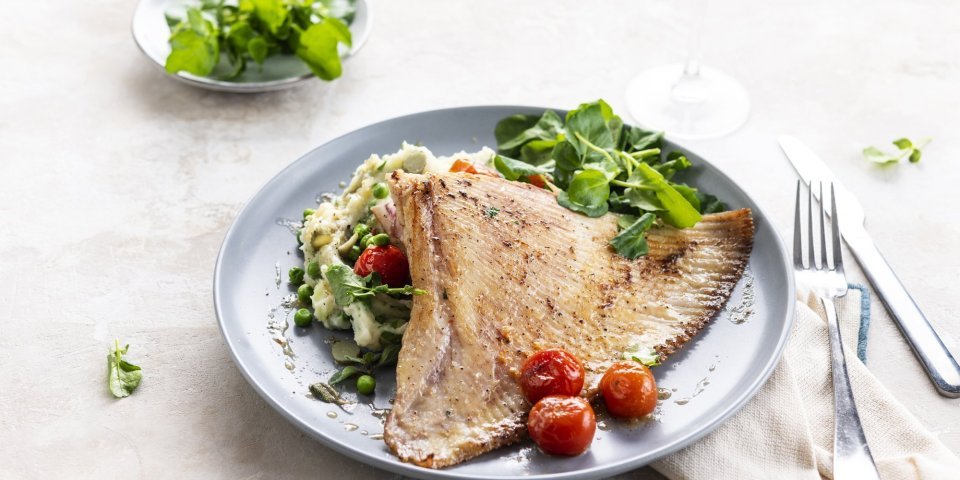 Een flinke schep puree is geschept op het bord. De puree heeft een leuk kleurtje door de erwten en rode kerstomaatjes. Hierop ligt de gebakken roggevleugel. We voegde nog enkele tomaatjes toe vanuit de oven en enkele blaadjes waterkers. 