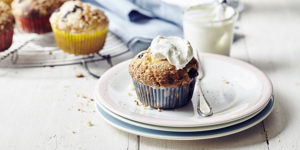 Er staan meerdere muffins op de tafel en eentje op een bordje dat al is afgewerkt met witte topping van slagroom of zure room.