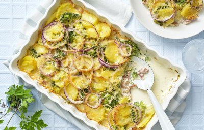 Een ovenschotel staat op tafel, waaruit al 1 hoek is uitgeschept op een bord. De ovenschotel bestaat uit krokante aardappelschijfjes, zoals een aardappelgratin, maar er is ook broccoli, rode ui en gerookte ham aan toegevoegd. Er staat een vaasje langs met een bloemetje en het tafelkleed is wit met ruitjes. 