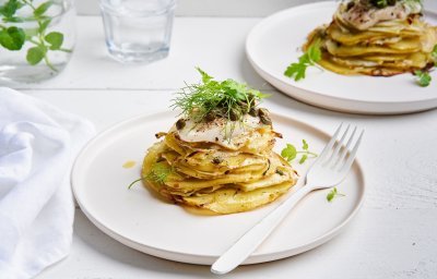 Op tafel staan twee borden, elk met een torentje van aardappelschijfjes, venkel en hierop een stukje pladijs. We werken af met kappertjes, olijfolie en wat venkelgroen. Er ligt een witte servette langs en een wit vork. 