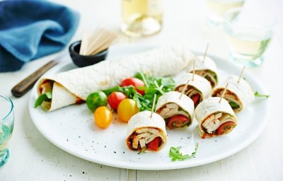 Op een bord ligt een rol wrap met groenten, wat kerstomaten en zes stukken wrap met een prikker in.
