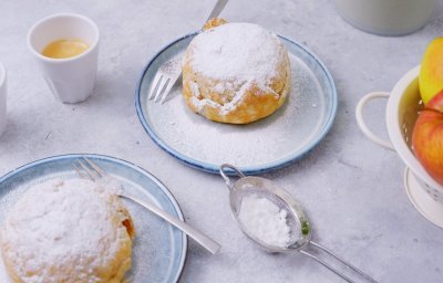 Heerlijke appelbollen staan klaar op tafel om toe te happen. Er staan twee kopjes kofie vij en verse jonagold appelen. De appelbollen zijn bestrooid met bloemsuiker. 