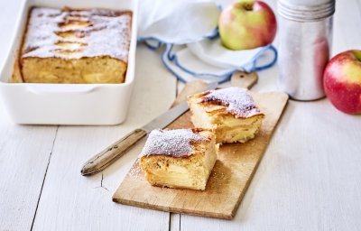 Een verse appelcake uit de oven, je reukt hem waarschijnlijk al. Er is een stukje uitgesneden en deze ligt opnieuw aangesneden op een houten plankje. Op tafel ligt een witte servette met blauwe randjes, nog enkele verse appels en de strooipot met bloemsuiker. 