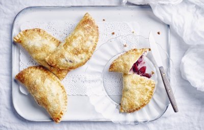 Er liggen 4 appelflappen klaargemaakt op een wit plateautje. Eén appelflap is opengesneden, door de combinatie te maken met blauwe bessen heeft de vulling mooi met appels een mooie rode kleur.