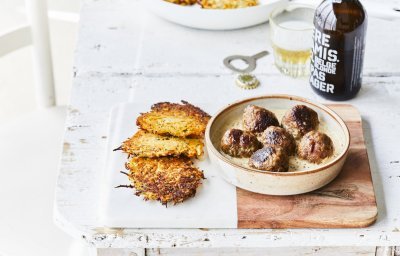 Gebakken balletjes liggen in een diep bord op de mosterdsaus, de krokante groenterösti's liggen er langs. Er staat een flesje bier op tafel met een klein glaasje.