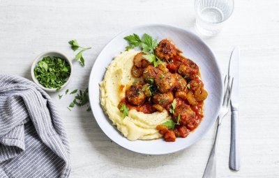 Een licht grijs bord staat op een witte tafel. Hierop is een schep smeuïge puree open geschept met daarbij de balletjes in tomatensaus. Het gerechtje is afgewerkt met wat verse peterselie. 