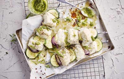 Klaargemaakte brochettes liggen in een grijze ovenschaal te blinken. De spiesjes bestaan uit stukjes heek, courgette en rode ui. Er staat een glazen potje naast met huisgemaakte dragonpesto.