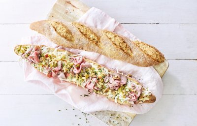 Een groot stokbrood ligt in de lengte opengesneden op een houten plank. Het broodje is belegd met een slaatje van knolselder, plukjes rosbief, stukjes appel en walnoten.