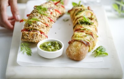 Twee broodjes liggen op een snijplank. Je ziet dat het twee versies zijn, nl een broodje met groene pesto, sneetjes ham en kaas. En dan nog een versie met roomkaas, paprika en rode ui. Extra pesto staat in een potje bij de broodjes, voor wie wat extra wil. Een vrouw zet de plateau of snijplank op tafel, haar handen zijn nog in beeld. 