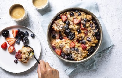 Homemade broodpudding met rabarber en zomerbessen staat op tafel. Een vrouwelijke gast schept haar een portie uit om volop te genieten van dit dessert met een kopje koffie. 