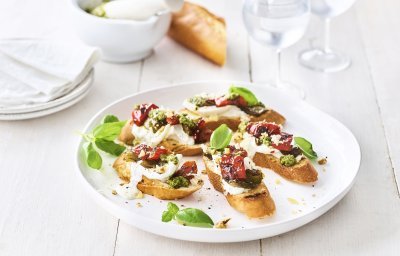 Op een houten, witte tafel staat een groot wit rond bord met hierop de bruschetta geserveerd. Ze zijn afgewerkt met extra blaadjes basilicum. Er staan ook kleine bordjes naast en de vijzel waarin de pesto is in gemaakt.