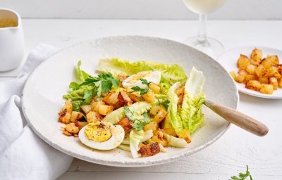 Op een houten witte tafel staat een diep bleek bord met hierin de caesarsalade. Hij is afgewerkt met de gekookte eitjes en goudgebakken croutons van aardappelblokjes. 