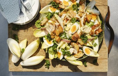 Op een houte snijplank staat een grote serveerschaal met hierin de caesarsalade geserveerd. Er staan enkele bordjes langs met bestek om onmiddelijk te starten. 